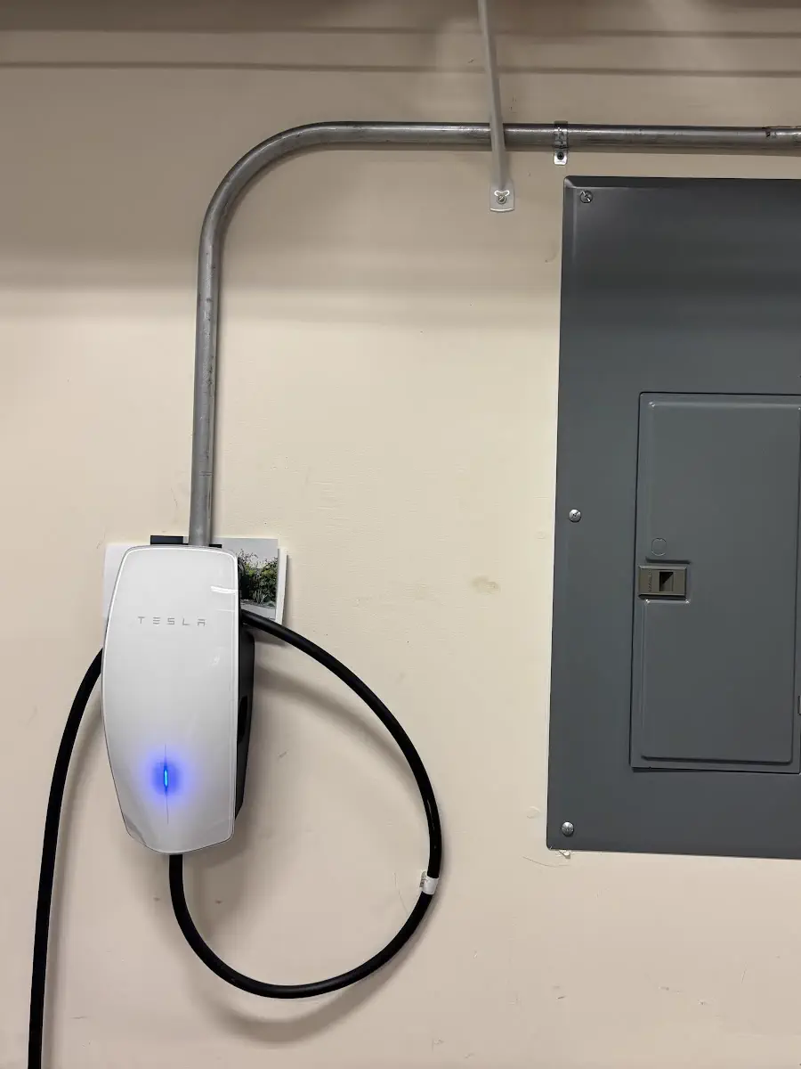 A Tesla wall charger in a garage, fed through conduit with a curved bend, beside two grey electrical panels
